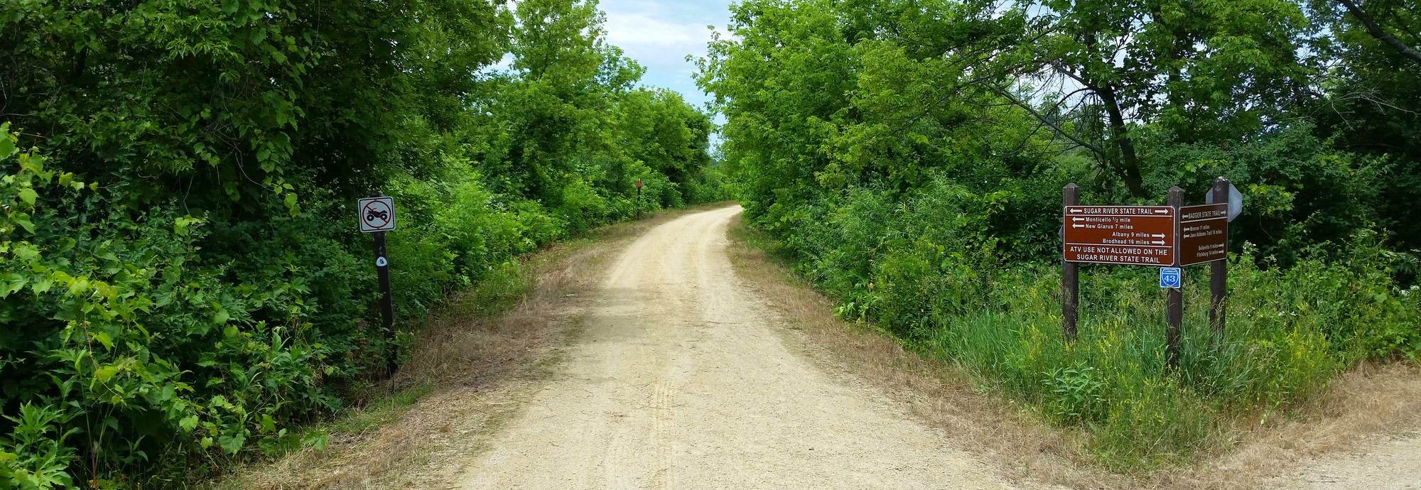 Crushed-limestone corridor of the Sugar River State Trail meeting a gravel crossing, dense summer canopy on both sides.