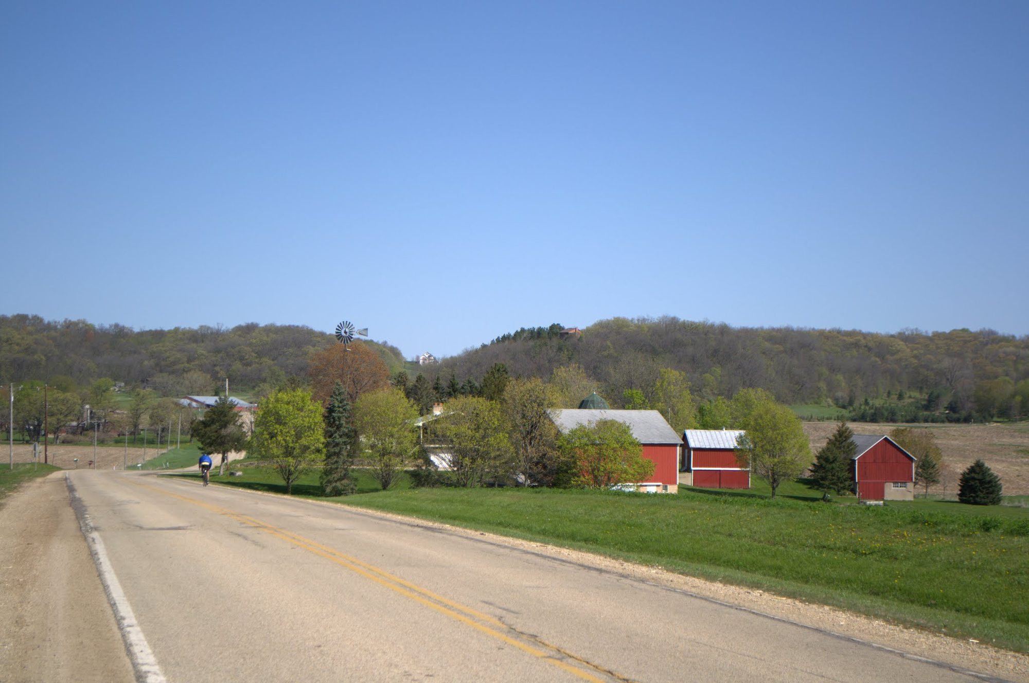 Rider on a two-lane county road past red barns, a windmill and wooded ridges in early spring.